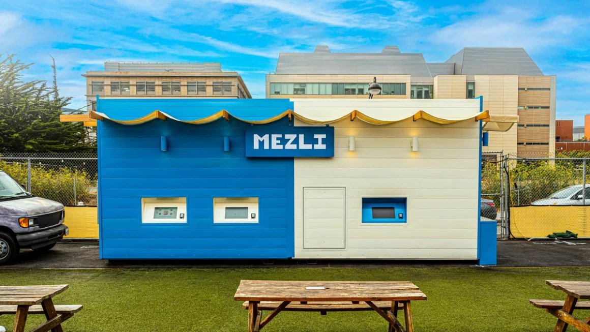 A container-sized restaurant in two colours, situated in a rural area with grass, picknick tables and buildings in the background.