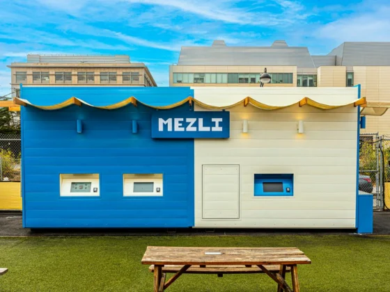 A container-sized restaurant in two colours, situated in a rural area with grass, picknick tables and buildings in the background.